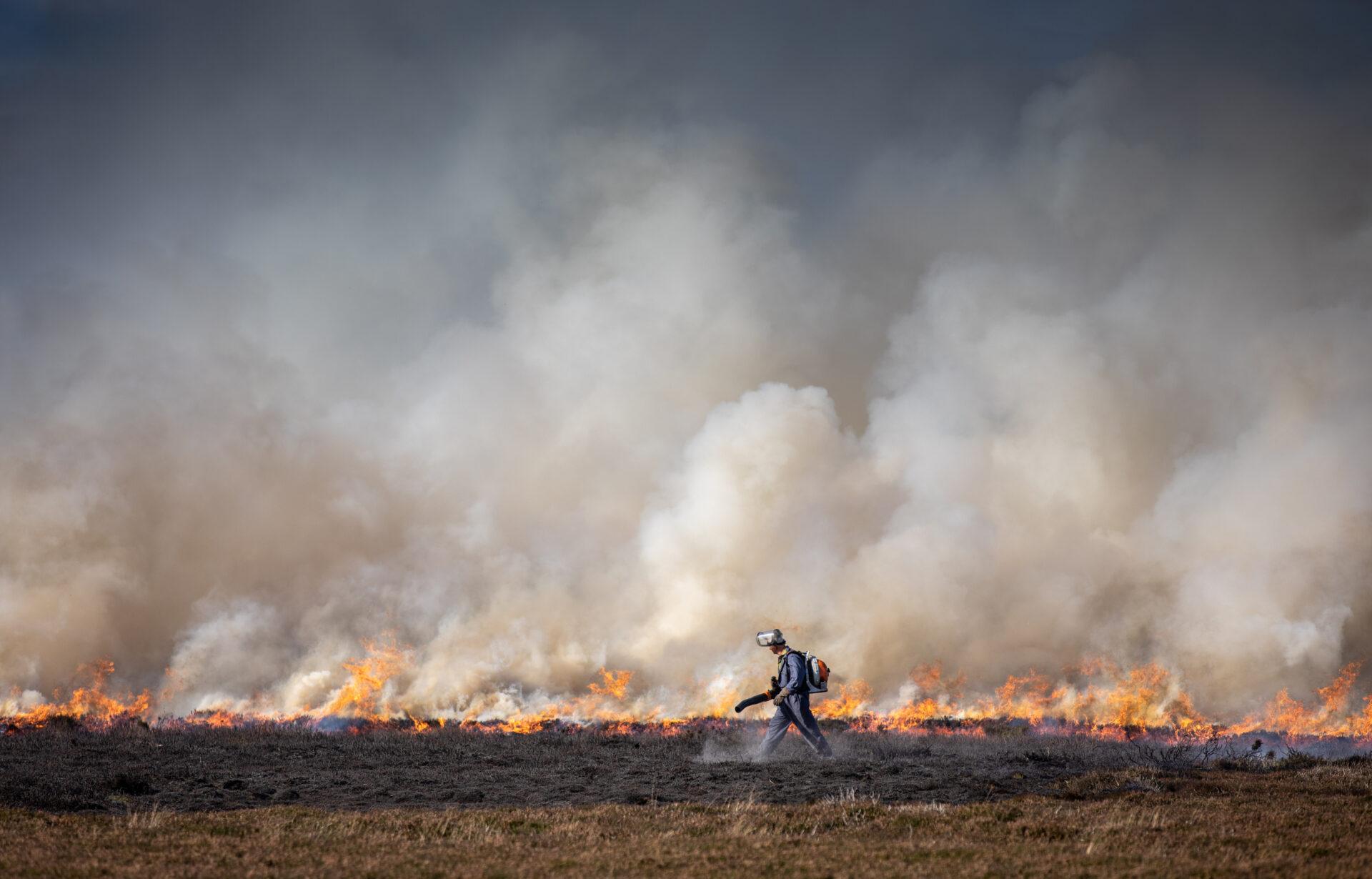Fire turns ground to ash and smoke clouds go into the air, as a man in fireproof gear walks carrying fire extinguisher.