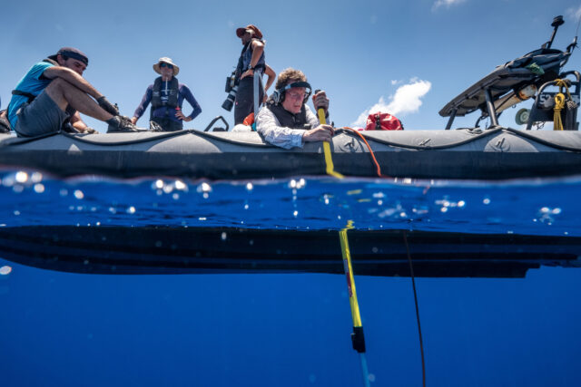 A person in an inflatable boat holds a long yellow pole with a cable attached into the water