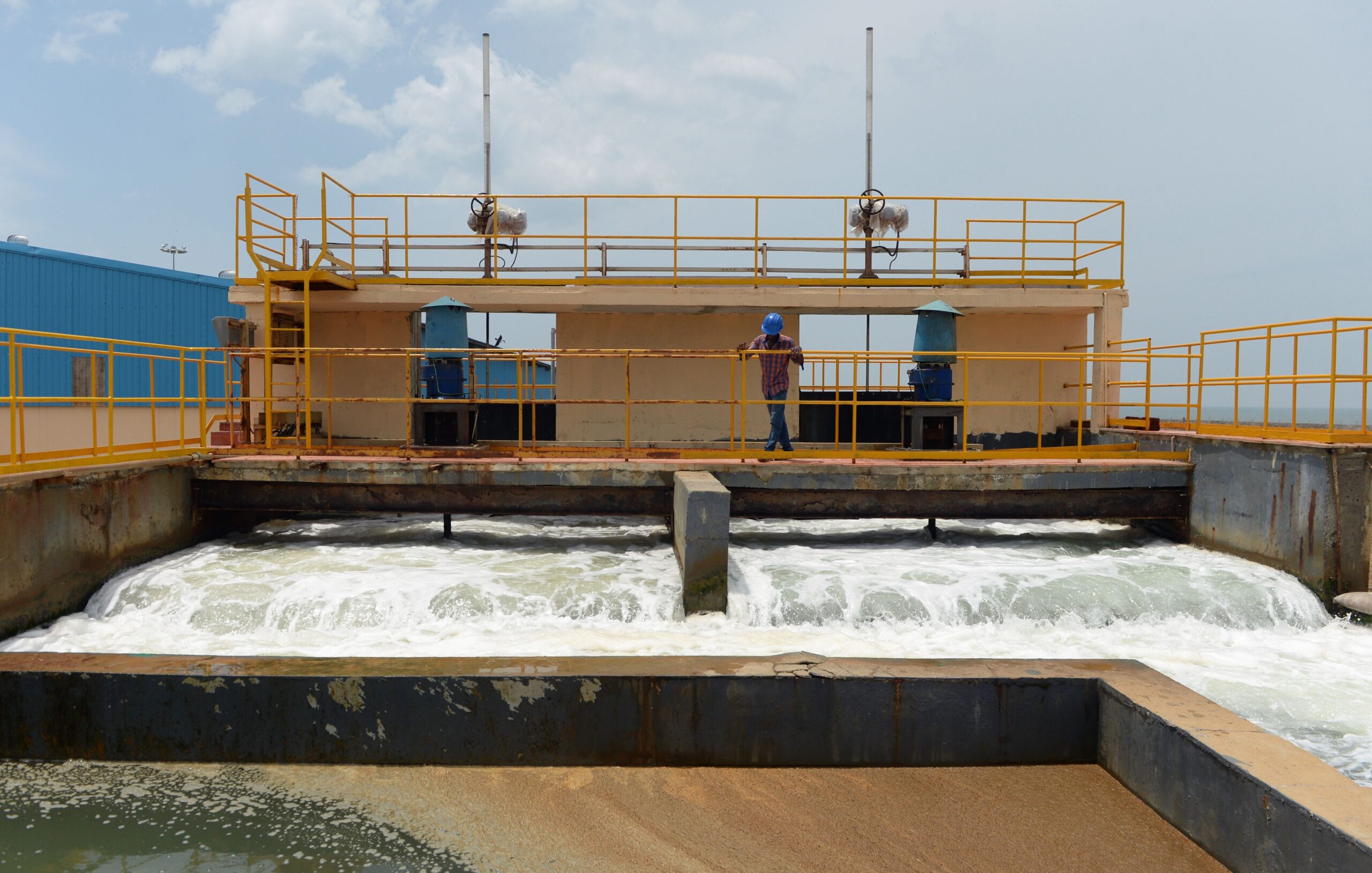 An industrial structure with water running through it. There is a bridge over the running water with a building, lined with yellow handrails and a man standing looking over the water flow.