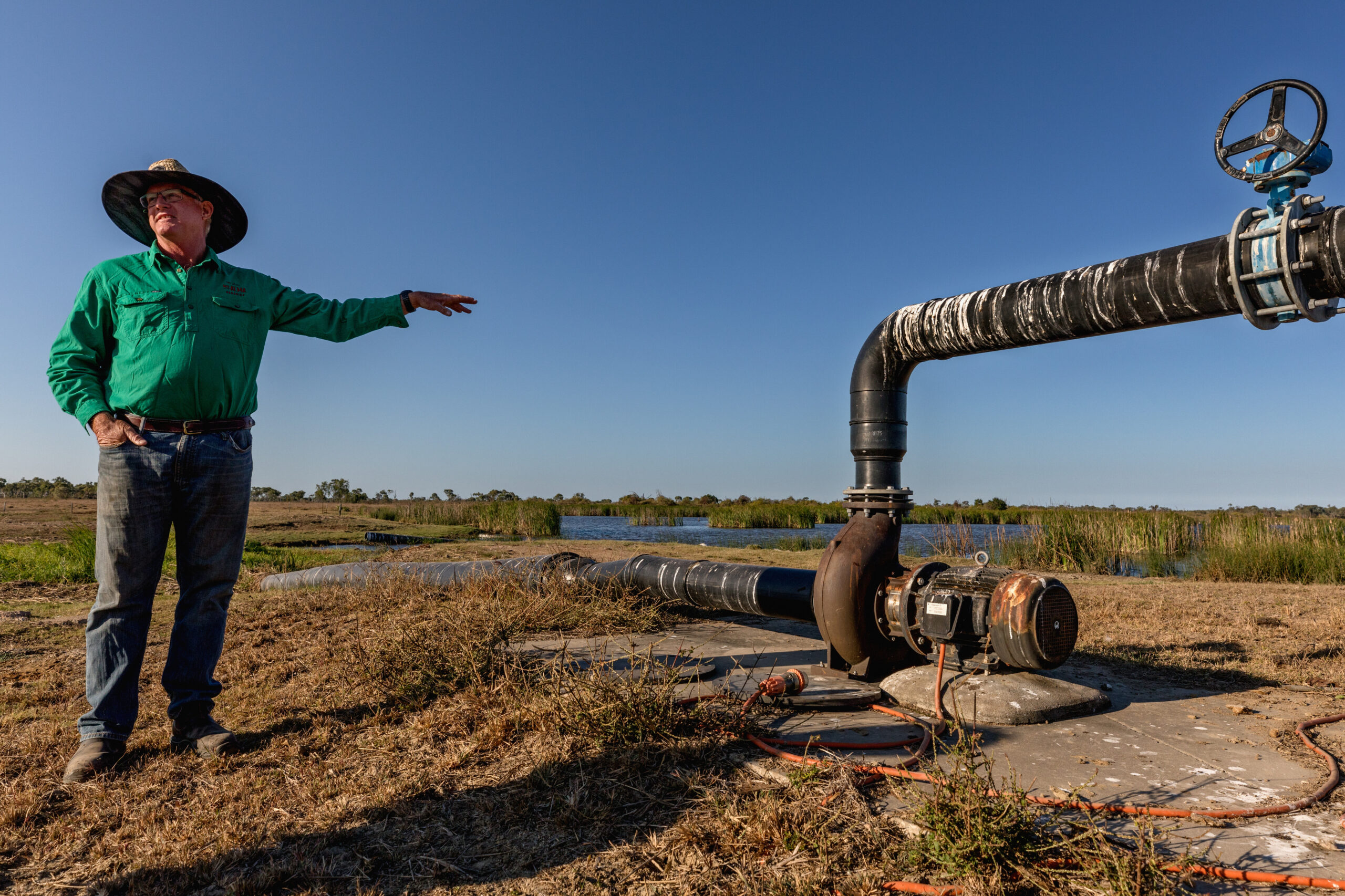 In a dry landscape with little vegetation, backed by a cloudless blue sky, a man stands in blue jeans a green shiry and a cowboy style hat, pointing over to his right, where a large metal pip with a raised section and tap appears to be pumping water to or from the land.
