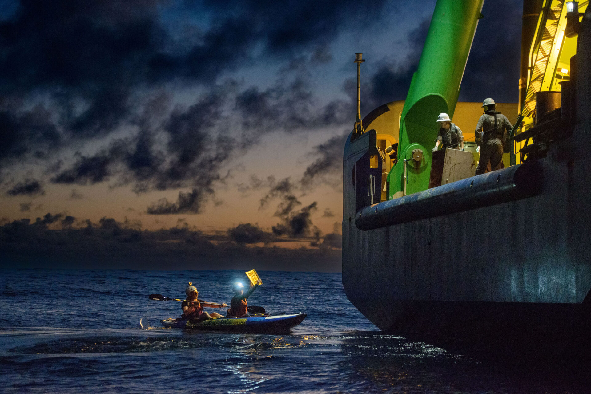 A dark scene with some final sunset light in the sky, with a large ship on the right towering over a small kayak with two activists; one is paddling and the other is holding a sign (illegible) up to the ship. Two people in white hard hats and boiler suits look down at the protesters from the side of the ship.