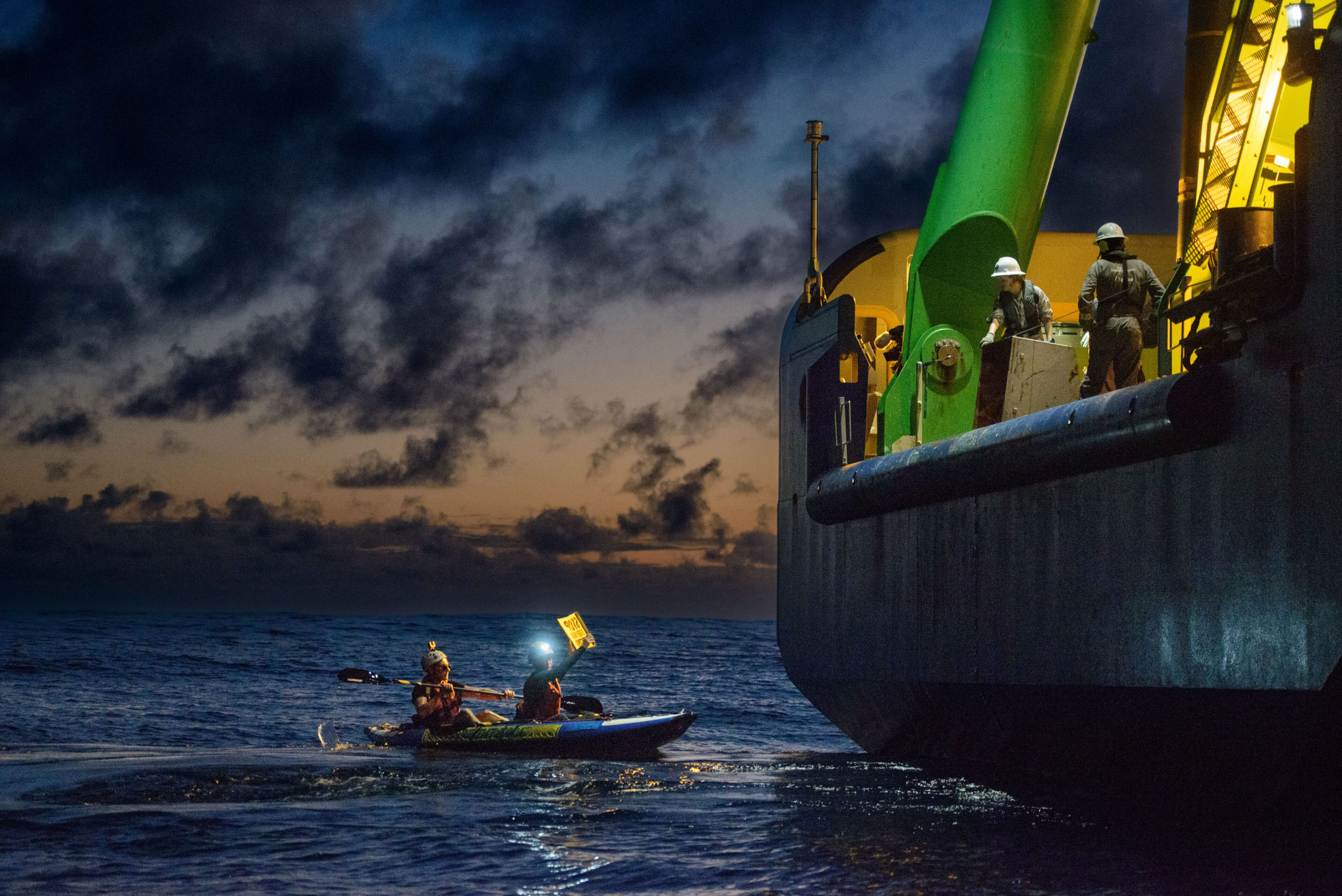 A dark scene with some final sunset light in the sky, with a large ship on the right towering over a small kayak with two activists; one is paddling and the other is holding a sign (illegible) up to the ship. Two people in white hard hats and boiler suits look down at the protesters from the side of the ship.
