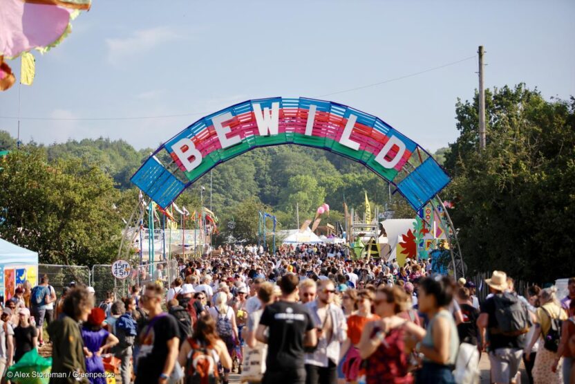 Crowds at a festival walk under a colourful wooden archway bearing the slogan 'be wild'.