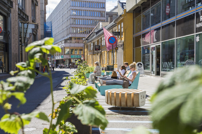 A number of people sunbathe on special street furniture sun-loungers (which have side tables next to them) in the middle of a wide, car-free street, surrounded by buildings. There are green leaves in the foreground.