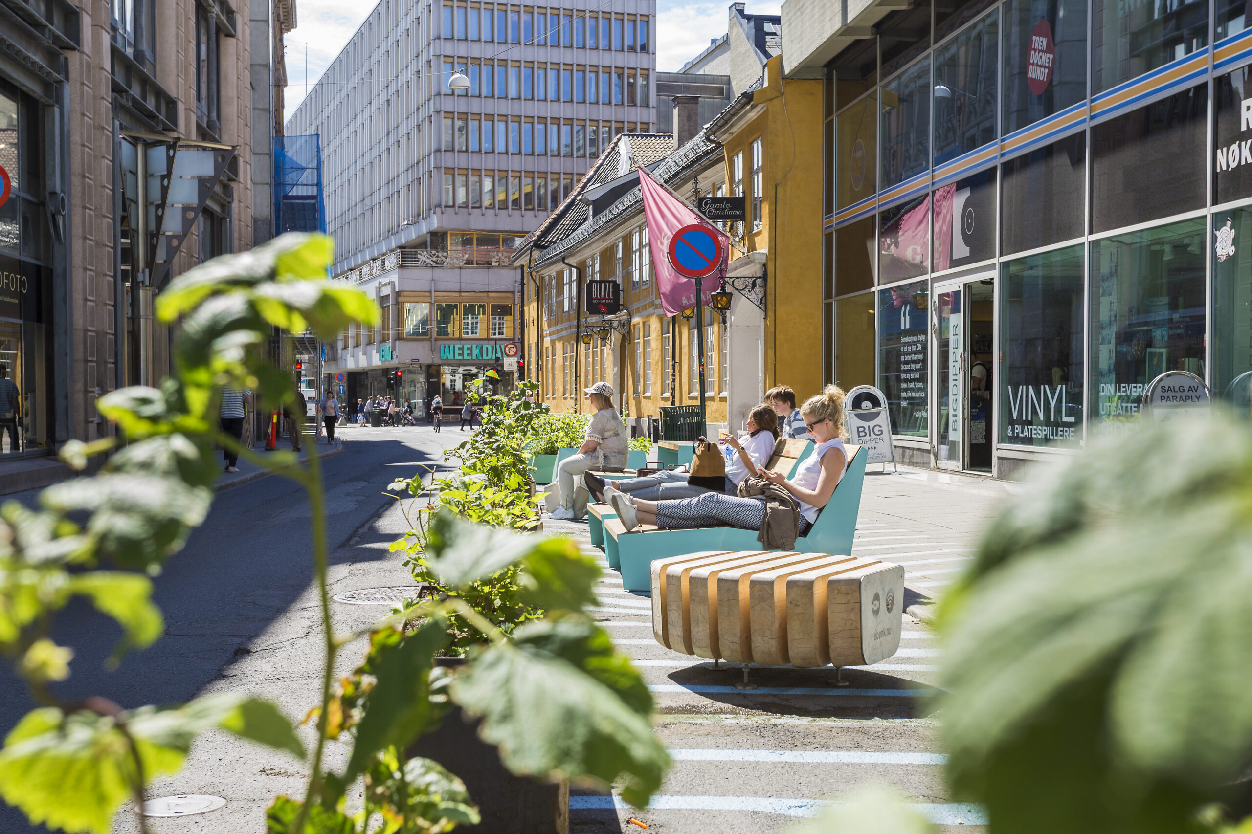 A number of people sunbathe on special street furniture sun-loungers (which have side tables next to them) in the middle of a wide, car-free street, surrounded by buildings. There are green leaves in the foreground.