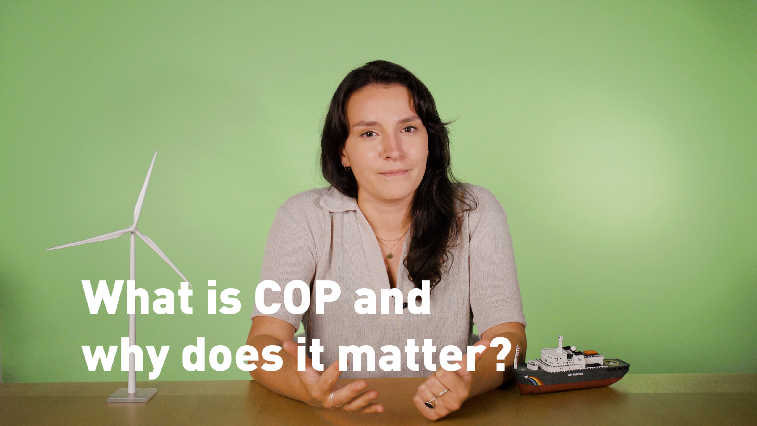 Video presenter sits at a desk decorated with a wind turbine and a model ship, against a plain green background. She looks into the camera with a half smile.