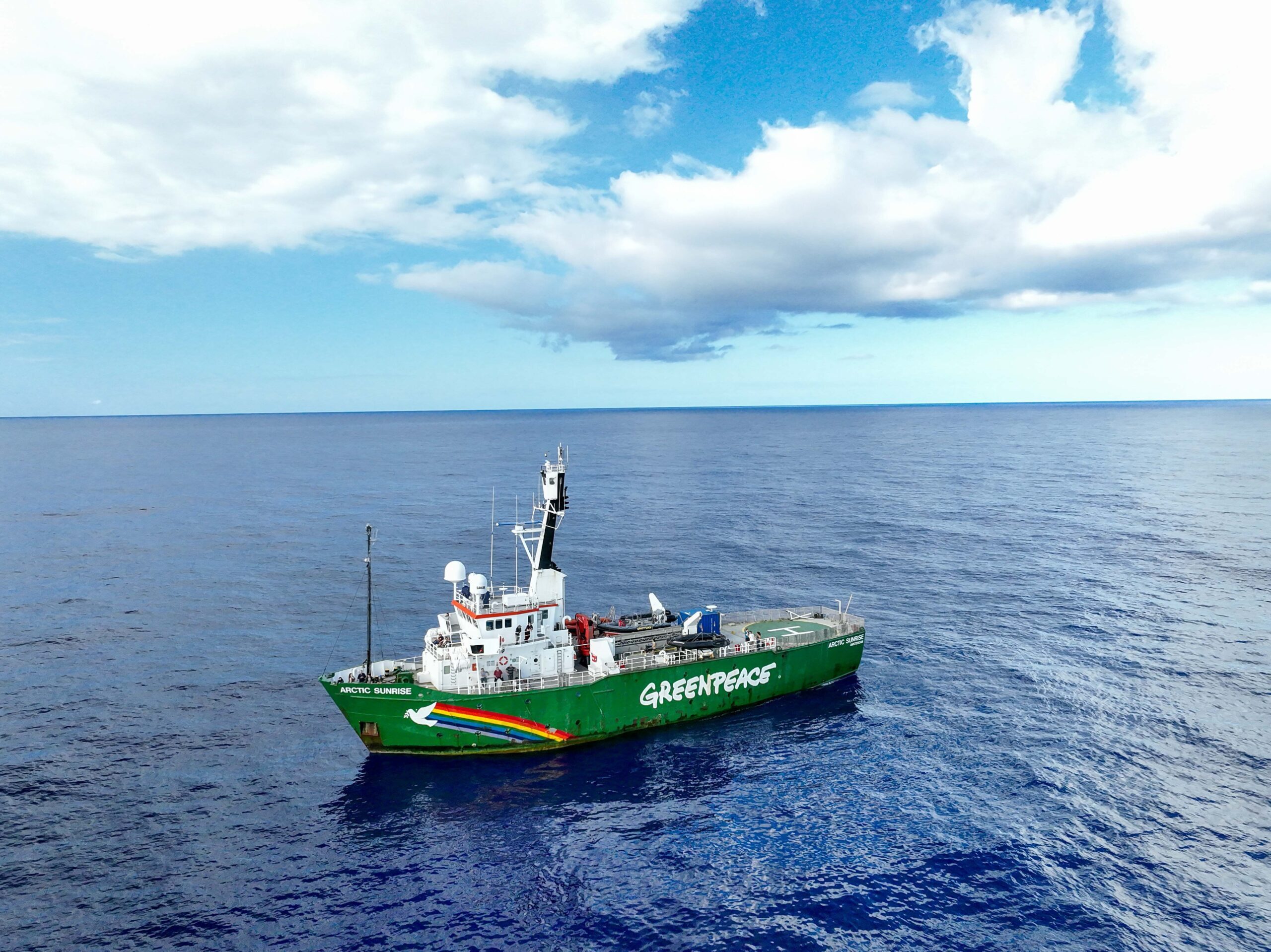 An image of a green and white ship in a calm sea, against a horizon and blue and cloudy sky in the distance. On the ship's green side is the word Greenpeace and a rainbow motif with a white bird