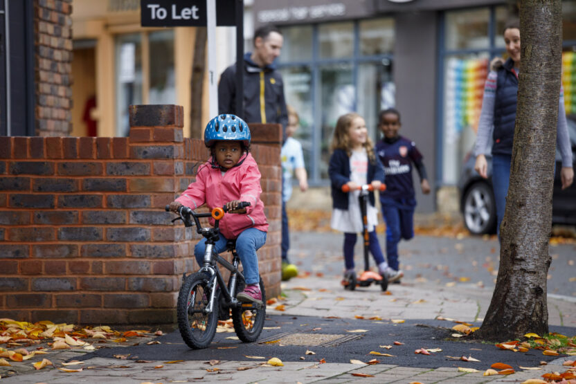 A young boy rides a bike along a residential street. A young girl on a scooter, a young boy running, and an adult walking are visible in the background.