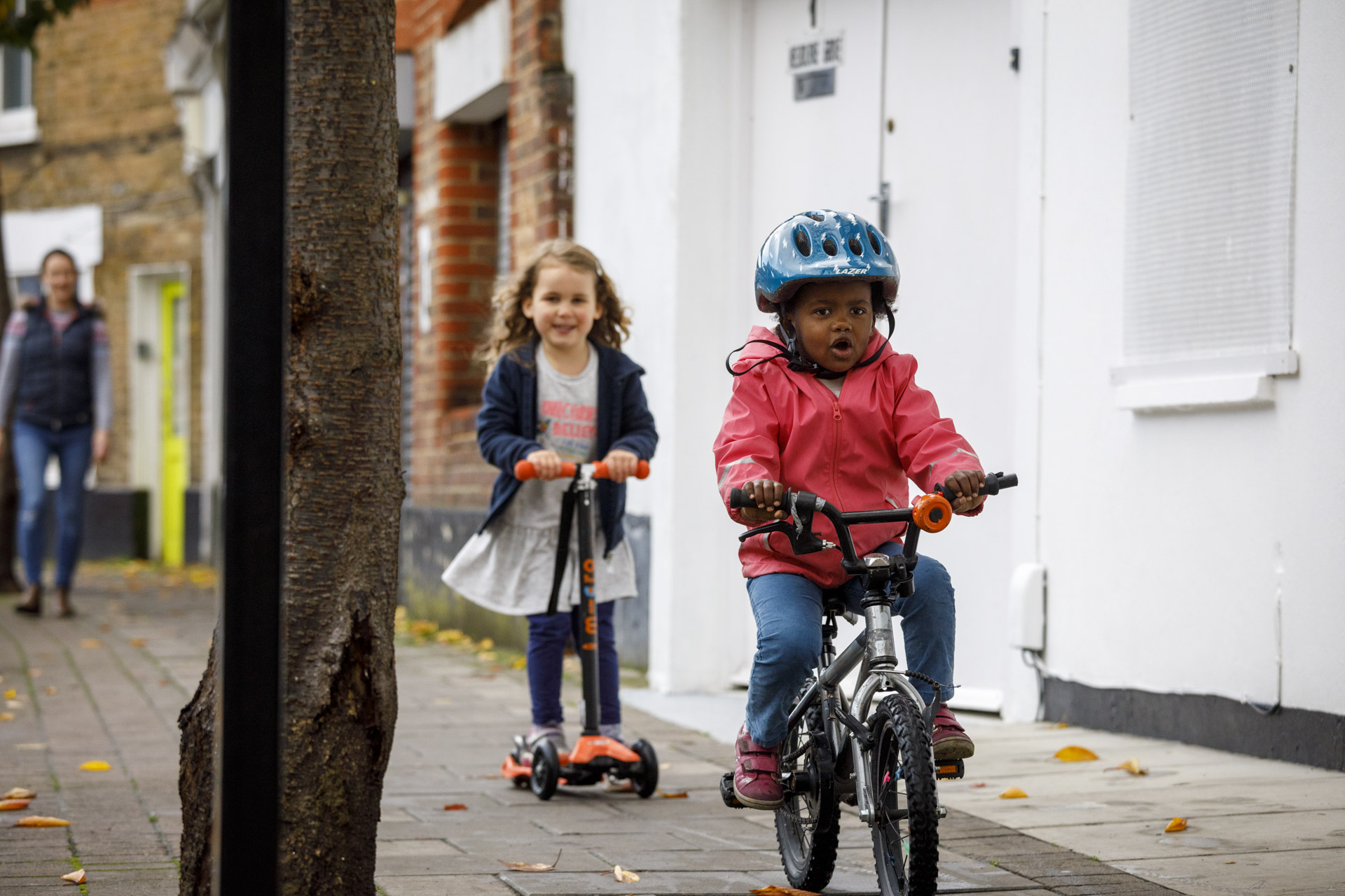 A young child on a bike rides towards the camera with a look of concentration on their face. Another child rides behind on a scooter.