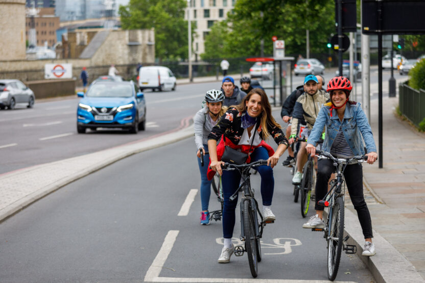 Two women on bikes smiling at the camera while waiting at a red light on a separated cycle lane. A queue of other riders is out of focus behind them.