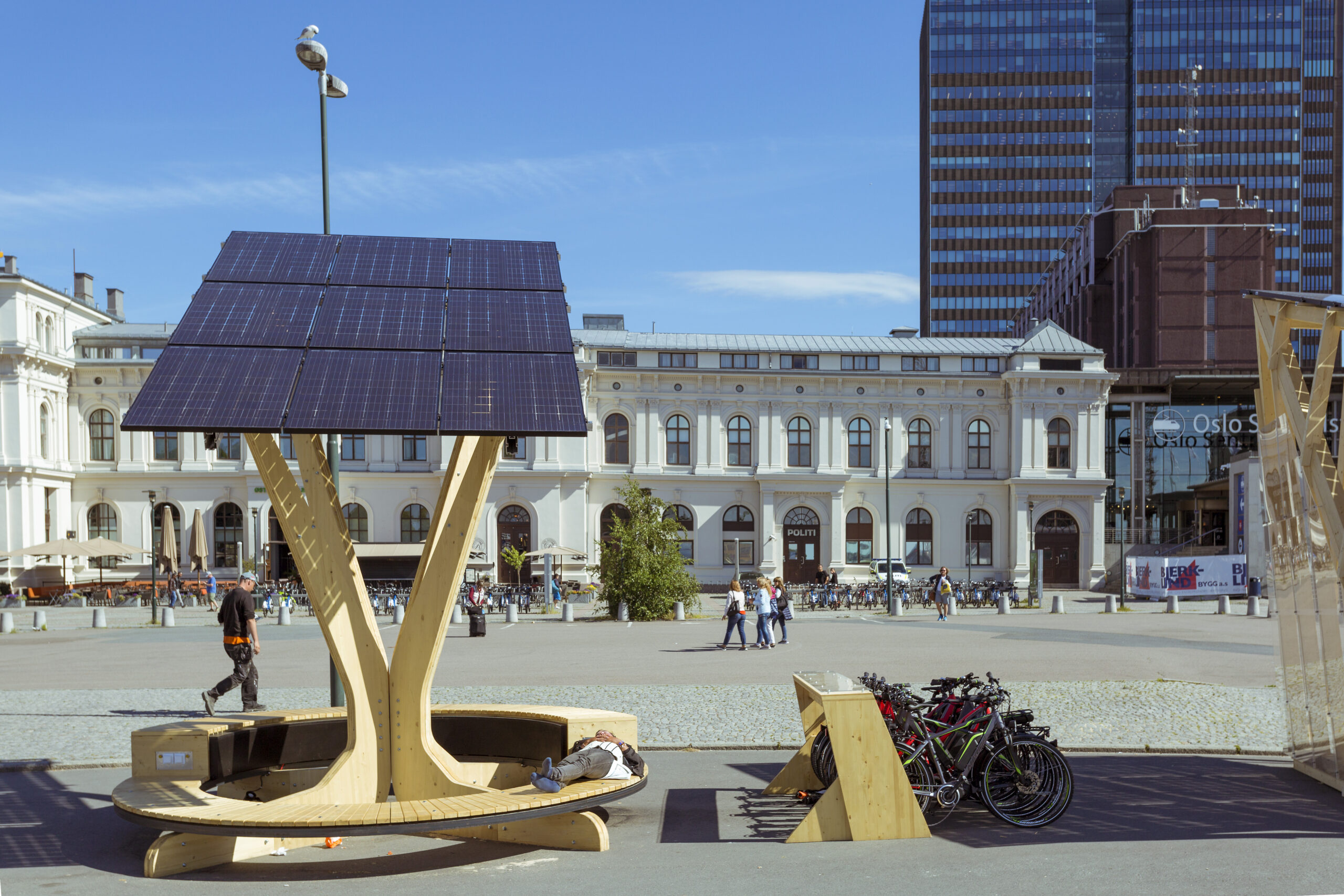 A bright sunny day in Oslo, a view of a wide square with official building in the background, and a bike docking station with a bench covered in a solar panel above it, on which someone is napping.