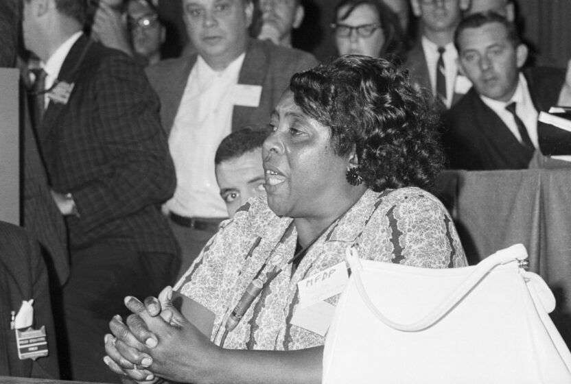 Fannie Lou Hammer is a Black woman. Her mouth is open mid-speech, as she sits and commands the attention of the people surrounding her.
