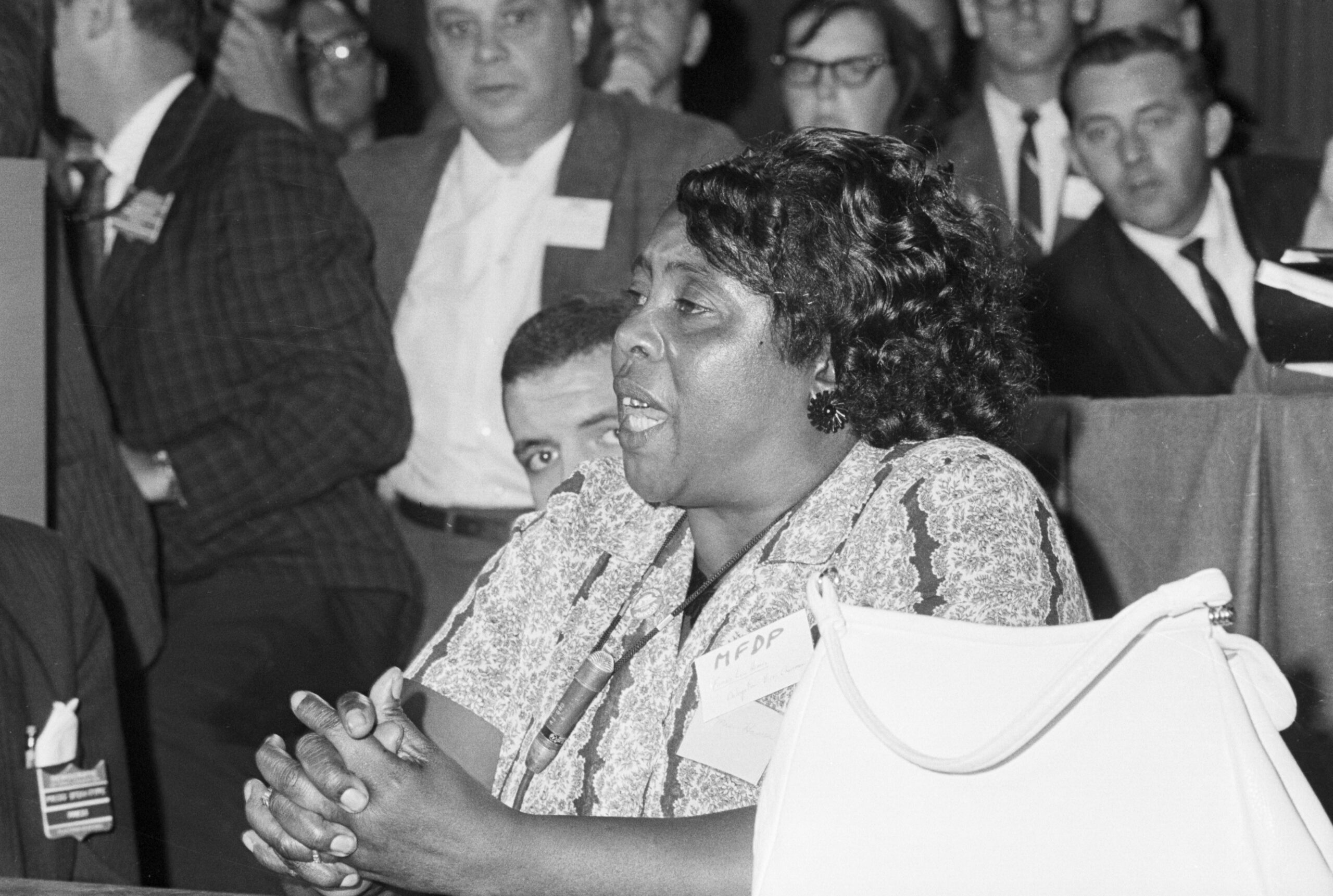 Fannie Lou Hammer is a Black woman. Her mouth is open mid-speech, as she sits and commands the attention of the people surrounding her.