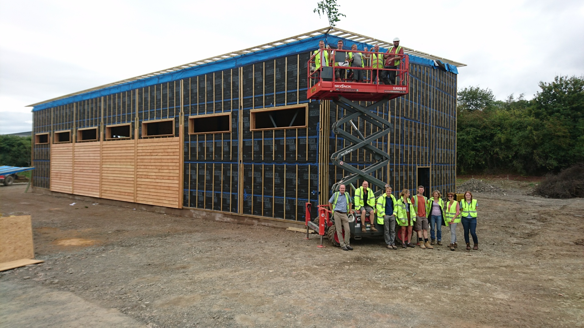 A building under construction with 14 people in front of it, 8 or so in front of an articulated lift and the others in the basket above them.