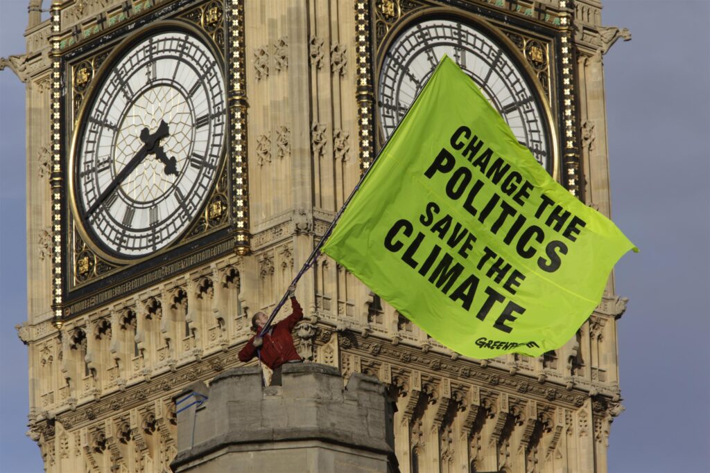 An activist stands on top of a stone tower waving a huge flag with the slogan "change the politics save the climate". Behind him, the iconic Big Ben clock looms just out of focus.