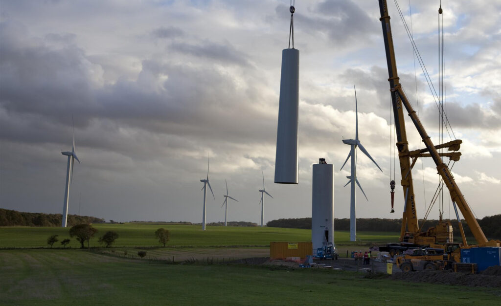 Giant cranes assemble the tower of a new wind turbine. A dramatic sunny sky and completed turbines can be seen in the background.