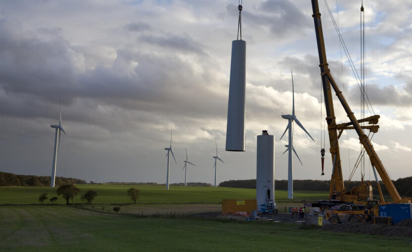 Giant cranes assemble the tower of a new wind turbine. A dramatic sunny sky and completed turbines can be seen in the background.
