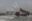 A truck carrying coal ash in a desolate and bleak landscape. There are coal chimneys in the distance with smoke going out into the atmosphere.