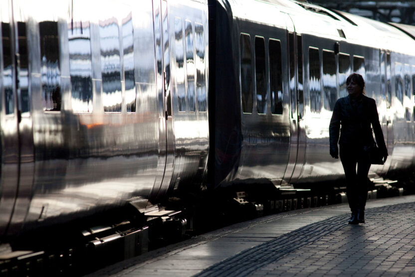 A passenger walking along a station platform is silhouetted against the sunlit carriages of a train.