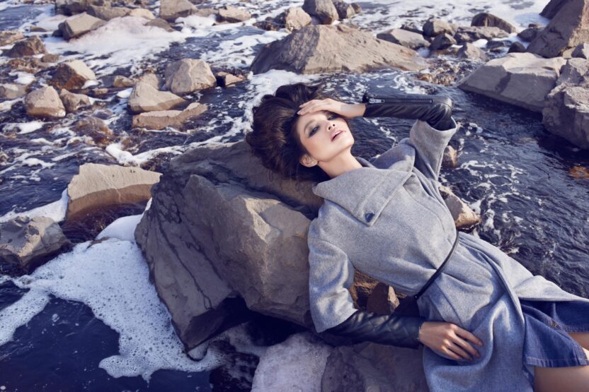 A model in a fashionably long grey coat lies on a rock in the middle of a polluted river