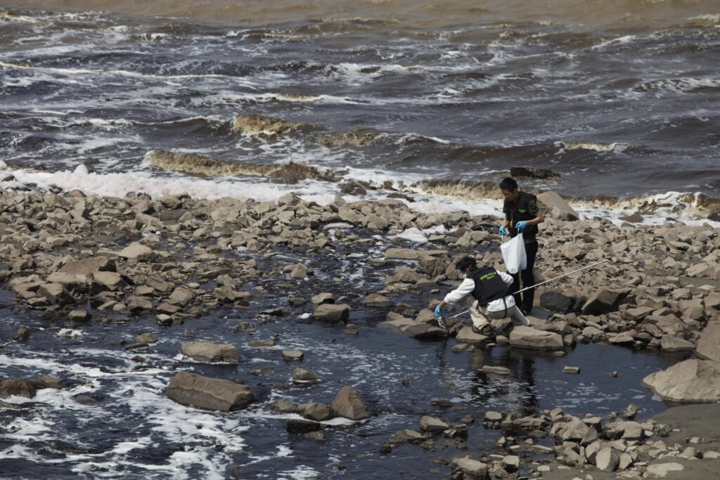 A rocky landscape with foamy, dirty water. Two people are collecting samples; one kneeling and the other one holding a bag