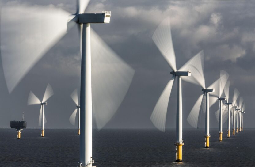 Offshore wind turbines lit by sun against a grey sky. Their blades are motion blurred.