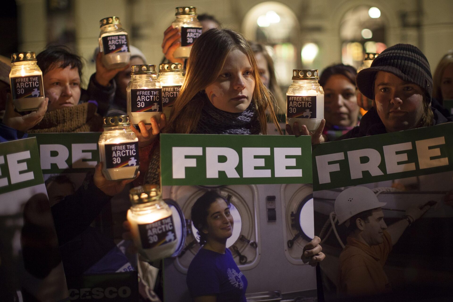 People at a candlelit vigil hold up 'Free the Arctic 30' signs with candles in jars.