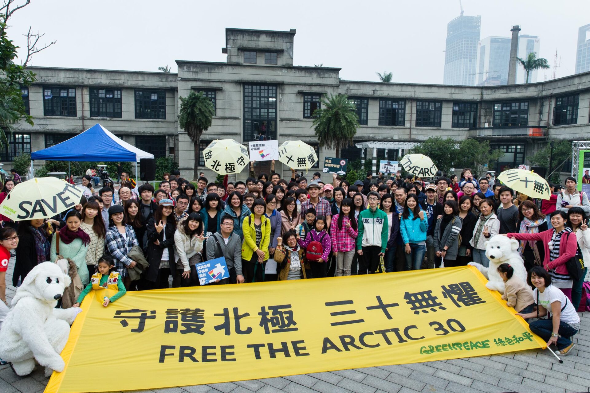 A crowd of protestors gathers round a banner that says 'Free the Arctic 30' in English with Chinese script above. There are also decorated umbrellas, placards and people in polar bear costumes.