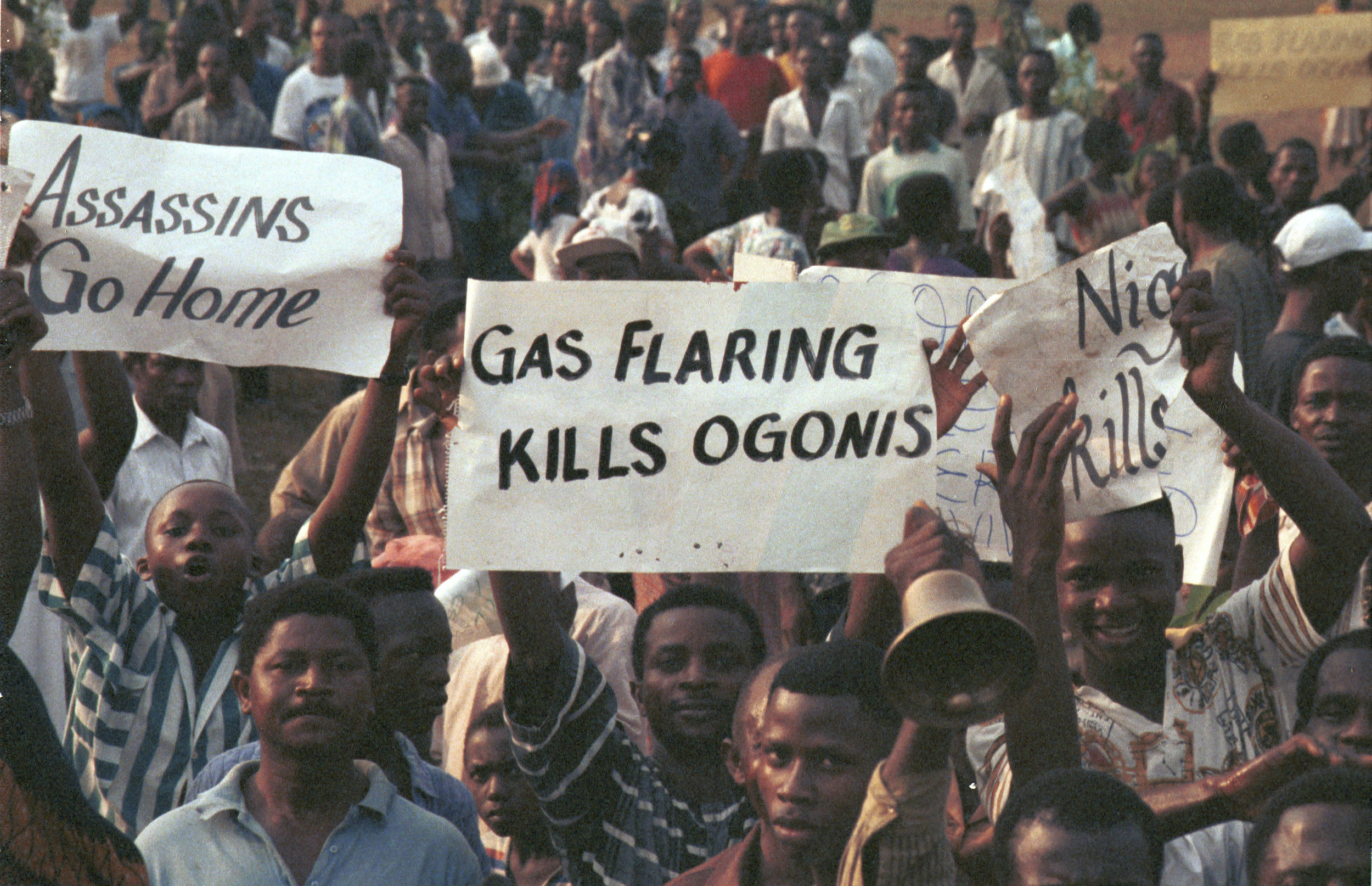 High angel view of demonstrating crowd at Ogoni Day demo, with banners: "Gas flaring kills Ogoni" and "Assassins go home". Ogoni Day demonstration, Nigeria, 1993