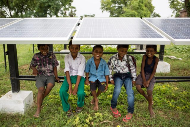 Children sit on the frame underneath a row of solar panels, smiling at the camera.