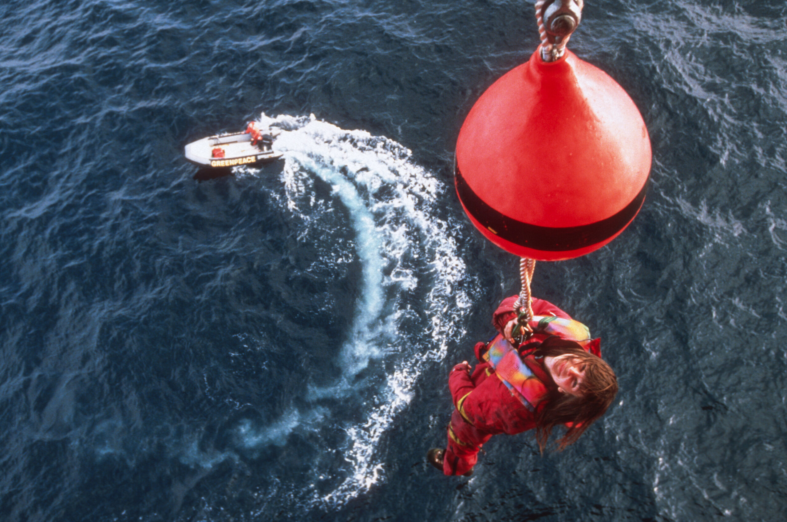 An activist smiles up into the camera while hanging on a rope high above the ocean. Below, a small boat is circling