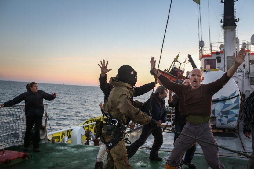 On the deck of a ship, a member of Greenpeace crew holds his hands up as he's accosted by a man in military uniform and a black balaclava.