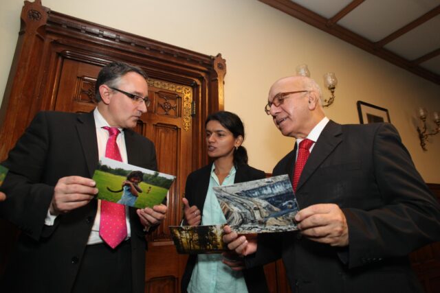 A volunteer wearing a Greenpeace branded tabard stands in front of a colourful Greenpeace banner speaking happily and confidently to an unseen audience.