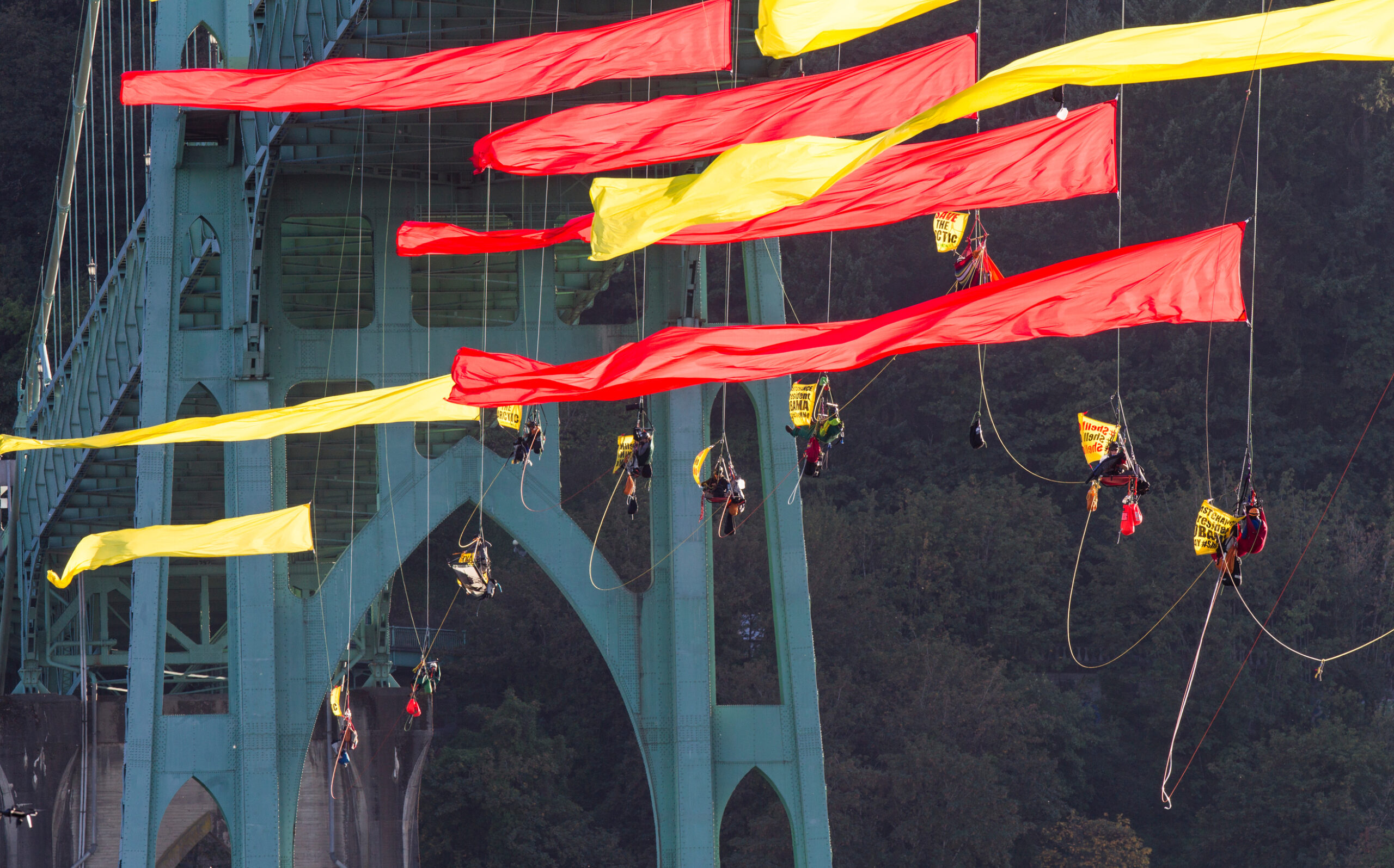 Activists in climbing gear hang from a giant river bridge. Huge red and yellow flags attached to their ropes fly proudly in the wind.