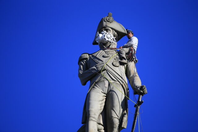 A Greenpeace activist sits on the shoulder of the giant Nelson statue in Trafalgar Square. The activist has put a giant respirator mask over the statue's face.
