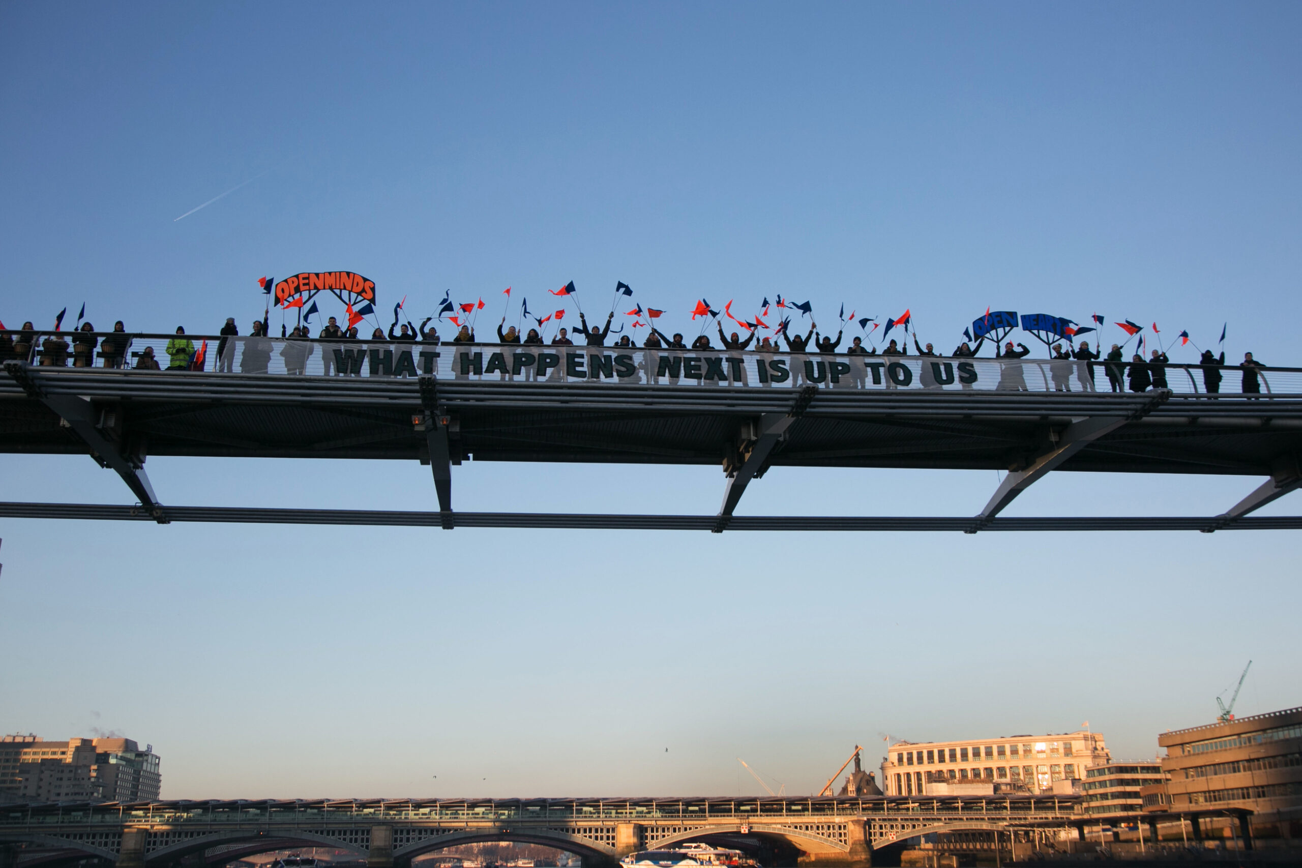 A close up of a part of a city bridge covered in people, against a blue sky, another bridge and and some buildings. The people on the bridge carry red and blue flags. A large banner central in the frame, reading "What comes next is up to us". smaller banners either side in red and blue read "Open minds" and "Open hearts"