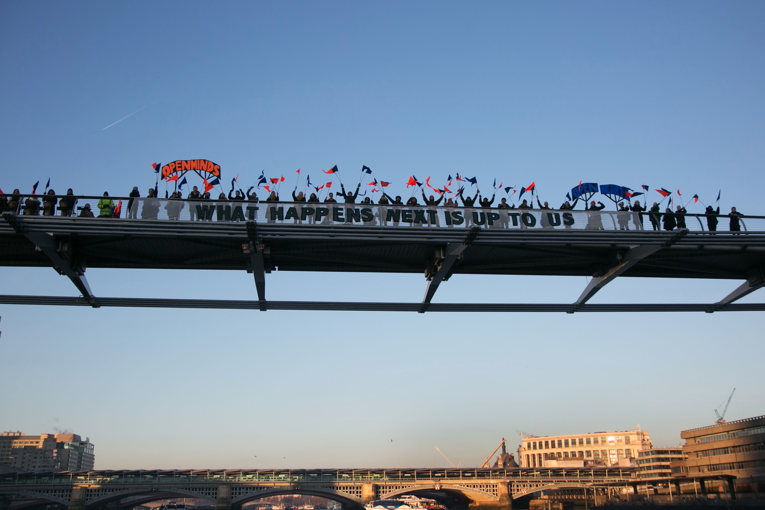 A close up of a part of a city bridge covered in people, against a blue sky, another bridge and and some buildings. The people on the bridge carry red and blue flags. A large banner central in the frame, reading 