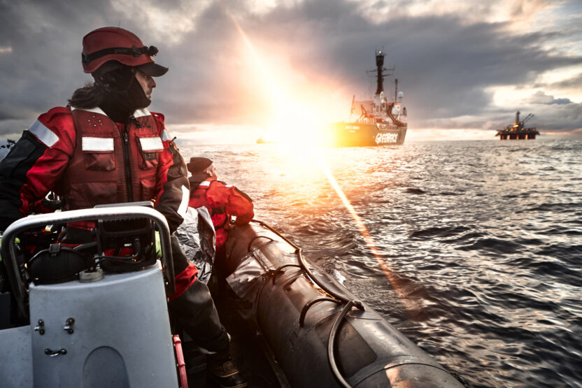 A greenpeace activist on a small inflatable motorboat looks back towards a Greenpeace ship in the sunset. An oil rig can be seen in the distance.