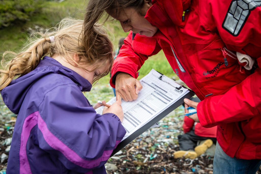 A young child dressed in a waterproof jacket examines a clipboard, while an adult looks on.