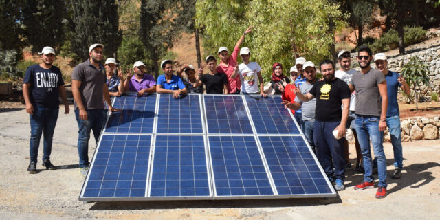 A group of trainees gathered around a small floor-mounted solar array. They're posing and smiling into the camera