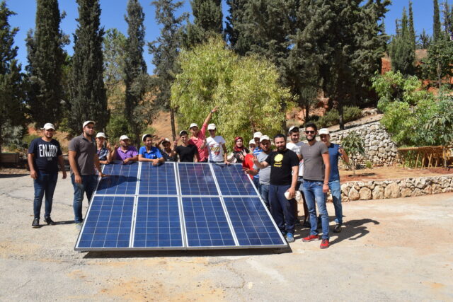 A group of people gathered around a small ground-mounted solar array, smiling at the camera.