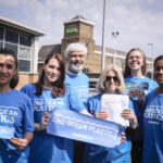 A group of six Greenpeace volunteers of different ages and ethnicities stand in the sunshine outside an ASDA supermarket. Smiling into the camera, they wear blue tshirts with 'End ocean plastics' slogans, and hold up campaign banners.