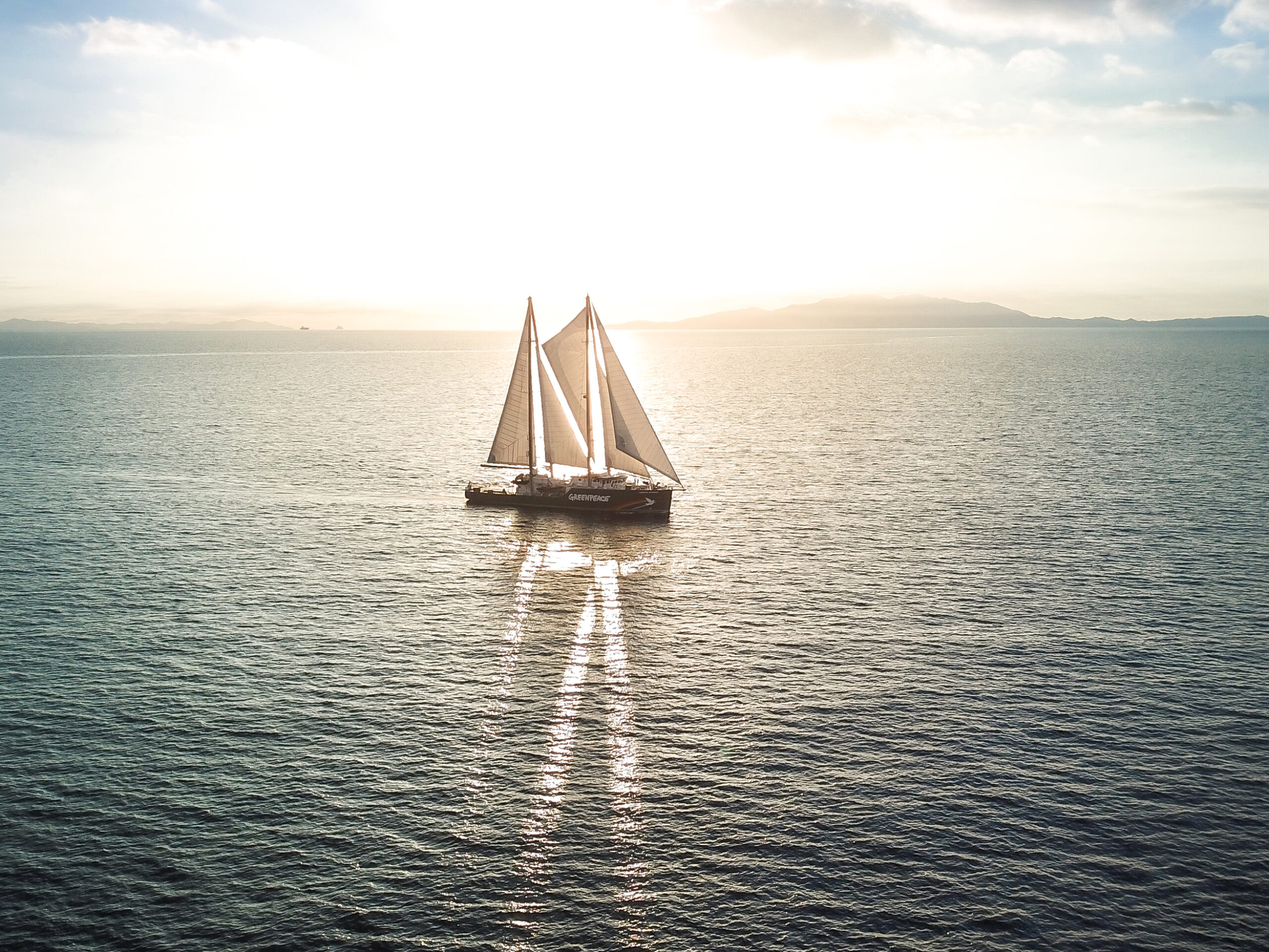 The Rainbow Warrior in open seas. The setting sun shines beautifully through its elegant white sails.