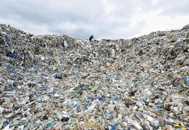A worker stands at the top of a giant mound of plastic waste