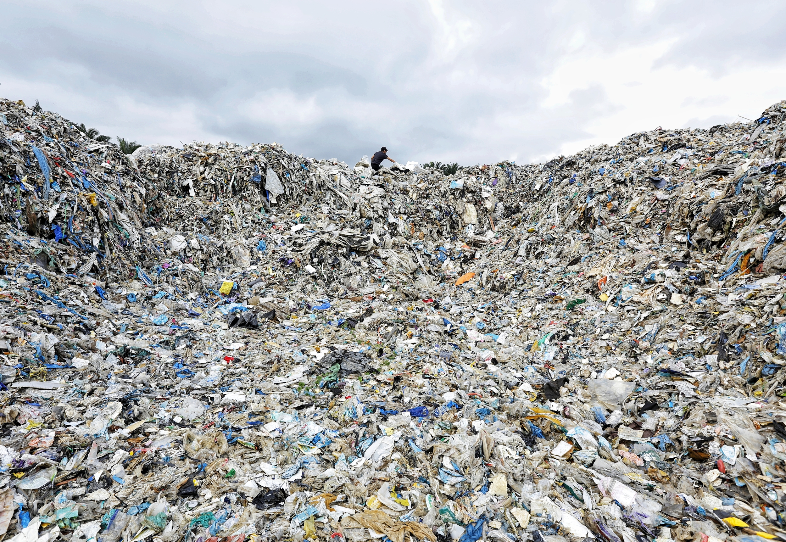 A worker stands at the top of a giant mound of plastic waste
