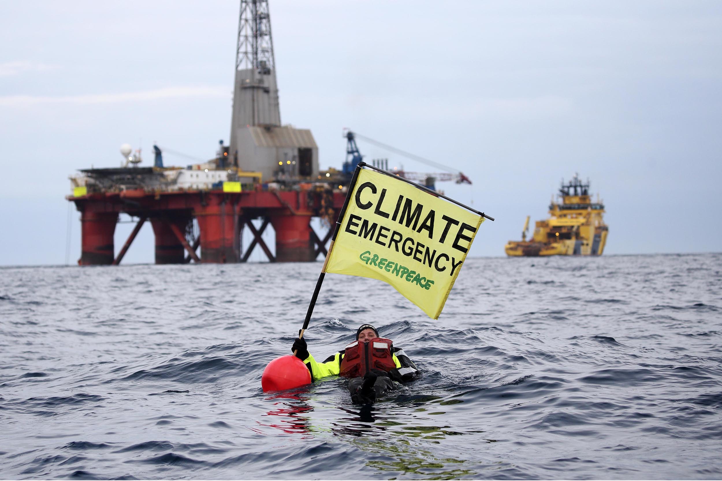 A swimmer floats in the sea, a short distance away from an oil rig. The flag they hold says 