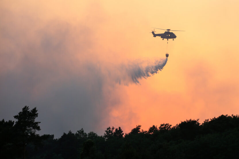 A helicopter silhouetted above a smokey forest against a burnt orange sky
