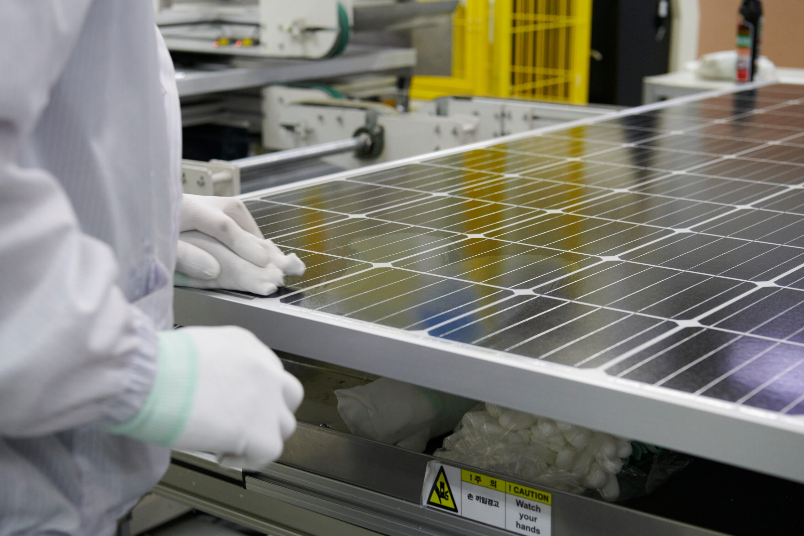 A close up of a solar panel in a factory with a person in shot in a white coat with white latex gloves touching one corner