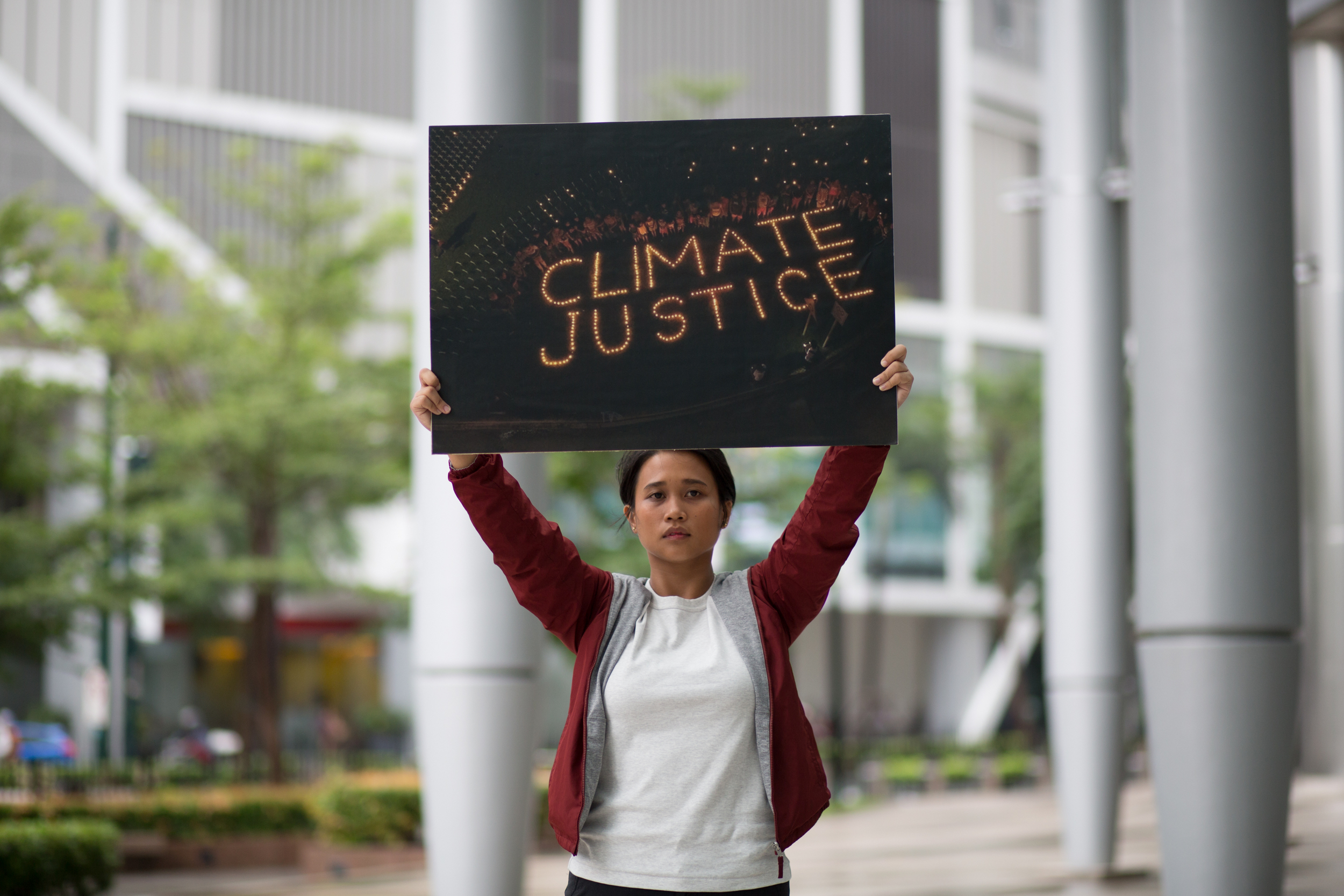A woman stands in front of a large building with a placard showing a photo where the words CLIMATE JUSTICE have been spelled out in candles, with a crown of people around them.