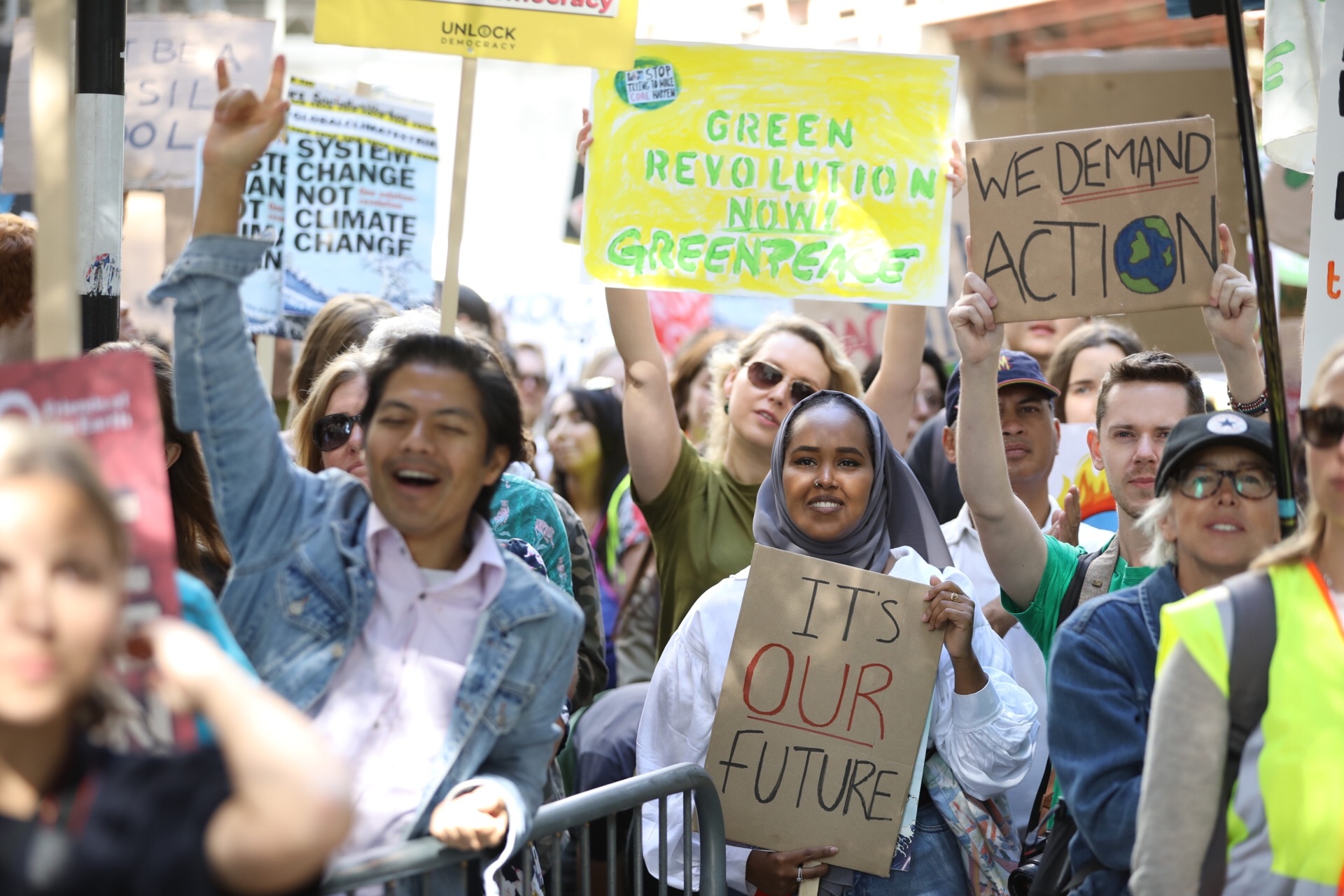 In bright sunshine, a smiling crowd of people hold up signs calling for a greener world. One protestor in the front row raises his fist in the air triumphantly.